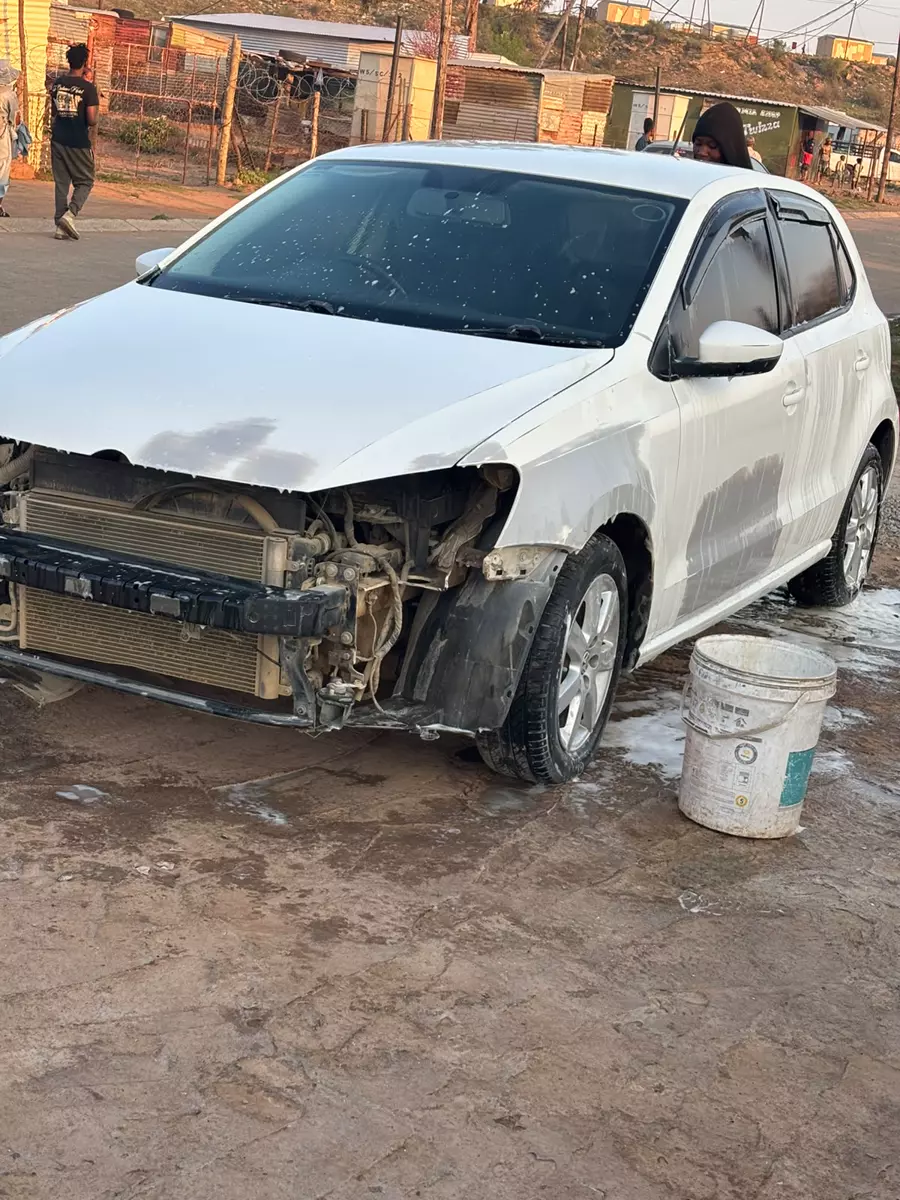 A damaged white car parked outside, undergoing a wash with a bucket nearby